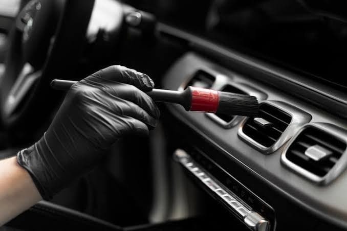 Gloved hand meticulously cleaning car dashboard air vents with a small detailing brush.